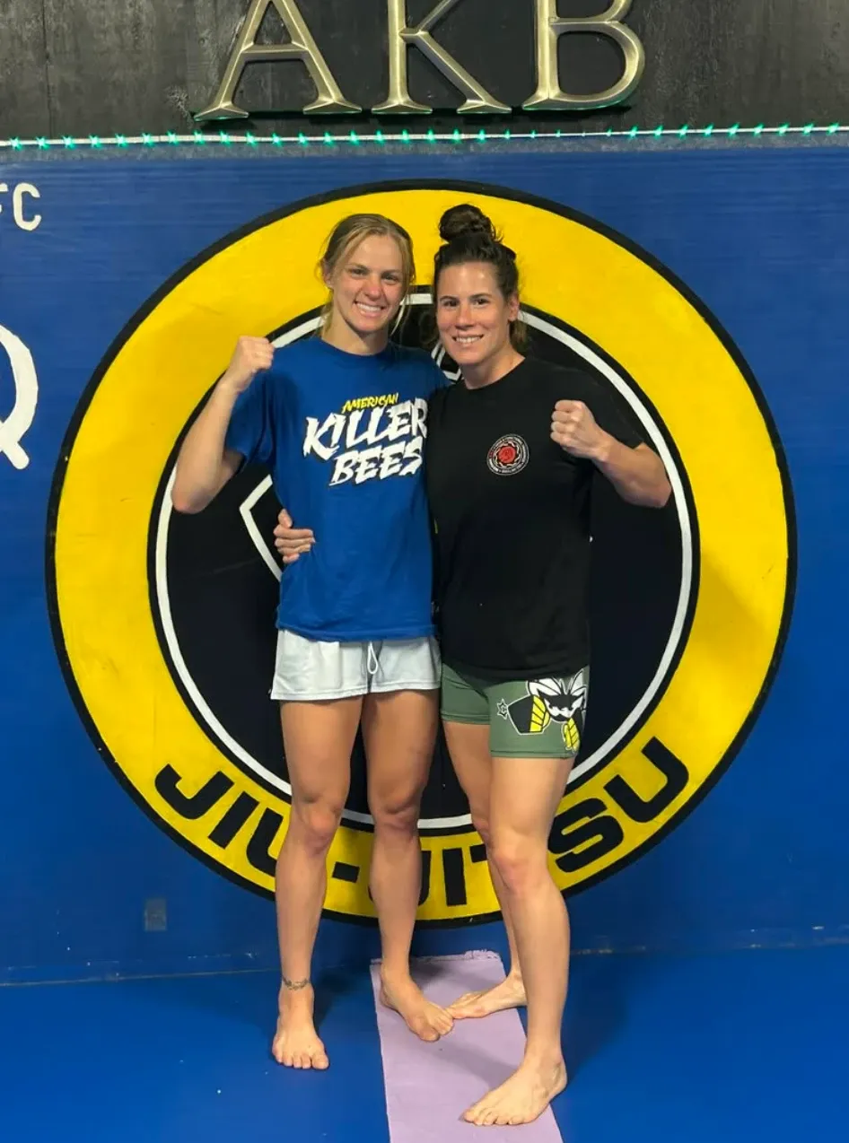 Victoria Leonardo Mason and Carli Judice with fists up in front of the American Killer Bees jiu-jitsu logo at the gym